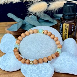 Könnte beinhalten: Ein Holzperlenarmband mit goldenen Akzenten und hellblauen Perlen. Das Armband wird auf weißen Steinen präsentiert, mit einer Flasche ätherischen Öls im Hintergrund. Das Armband besteht aus kleinen, runden, hellbraunen Holzperlen.