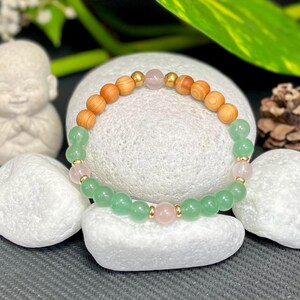 Könnte beinhalten: Ein Armband mit Holz-, grünen und rosafarbenen Edelsteinen. Das Armband besteht aus runden Holzperlen, grünen Aventurinperlen und Rosenquarzperlen, die mit goldfarbenen Abstandshaltern aufgereiht sind.