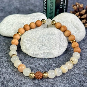 Könnte beinhalten: Ein Armband mit hellbraunen Holzperlen, cremefarbenen und pfirsichfarbenen Edelsteinperlen und goldfarbenen Metallakzenten. Das Armband wird auf einer grauen Oberfläche mit weißen Steinen und einer Flasche ätherischen Öls im Hintergrund präsentiert.