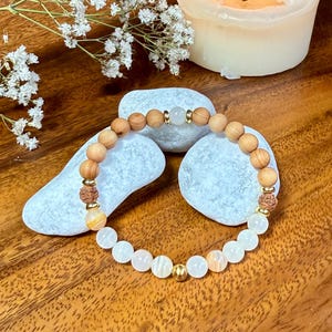 Könnte beinhalten: Ein Armband mit hellbraunen Holzperlen, weißen und pfirsichfarbenen Edelsteinperlen und goldfarbenen Metallakzenten. Das Armband wird auf einer Holzoberfläche mit weißen Steinen und einer brennenden Kerze im Hintergrund präsentiert.