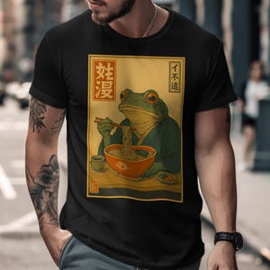 May include: Black t-shirt featuring a frog illustration. The frog is depicted in a traditional Japanese style, wearing a robe and eating ramen. The image includes Japanese text and a bowl of noodles.