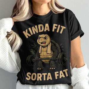 Op de afbeelding: Zwart T-shirt met de gouden tekst "KINDA FIT SORTA FAT". Het ontwerp toont een cartoon schildpad die gewichten heft. De schildpad heeft een vastberaden uitdrukking en draagt een handdoek.