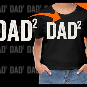 May include: Black t-shirt with the white text "DAD²" and an orange arrow pointing to the right. The shirt is worn by a person in blue jeans. The background has a repeating blurred pattern of the same text.