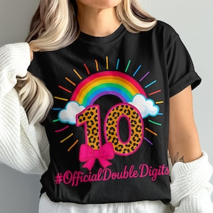 Op de afbeelding: Zwart T-shirt met een kleurrijke regenboog, wolken en het nummer 10 in een luipaardprint. Een roze strik staat onder het nummer 10, met de tekst "#OfficialDoubleDigits" onderaan.