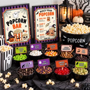 Op de afbeelding: Een Halloween popcornbar met twee ingelijste borden, een zwarte ketel gevuld met popcorn en verschillende kleine zwarte schaaltjes met diverse toppings. Elk schaaltje heeft een label met namen zoals Spider Eggs en Ghost Puffs.