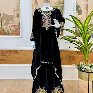 Könnte beinhalten: Ein schwarzes Samtkleid mit langen Ärmeln und aufwendiger Stickerei in Rot, Gold und Weiß. Das Kleid hat detaillierte Muster auf dem Oberteil, den Ärmeln und dem Saum, mit einer fließenden Silhouette und zarten weißen Verzierungen.