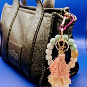 May include: A black leather handbag with a decorative keychain attached. The keychain features a pink carabiner, wooden beads, and a layered tassel in shades of peach and cream. The background is a solid blue.