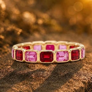 Könnte beinhalten: Ein goldener Eternity-Ring mit einer durchgehenden Reihe abwechselnd im Smaragdschliff geschliffener roter und rosa Edelsteine. Der Ring zeigt ein wiederkehrendes Muster aus leuchtend karmesinroten und zart rosafarbenen Steinen in einer Goldfassung.
