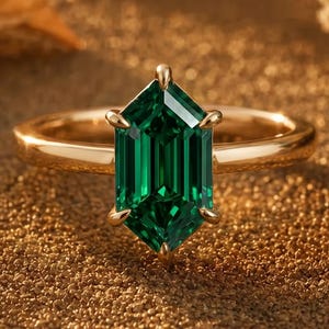 May include: A gold ring featuring a central hexagonal-cut emerald green gemstone held by six prongs. The band is smooth and polished, showcasing a warm metallic lustre that complements the deep green hue of the faceted stone in the centre.