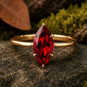 May include: A gold ring featuring a marquise-cut, deep red gemstone. The gemstone is held in place by four prongs. The ring is set against a natural, textured background with hints of green and brown.