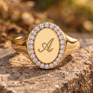 May include: A gold signet ring featuring an oval face with a polished centre engraved with the letter A. The oval face is surrounded by a halo of small, clear, round-cut stones set in a gold band that tapers towards the back of the ring.