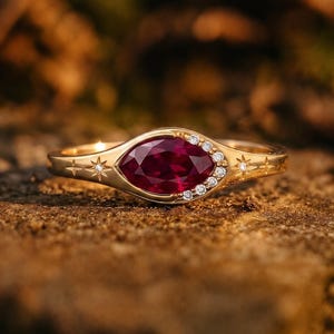 May include: A gold ring featuring a central marquise-cut deep red gemstone. The band is accented with small round white stones along one side of the setting and engraved star motifs on the shank, creating a celestial-inspired jewelry piece.