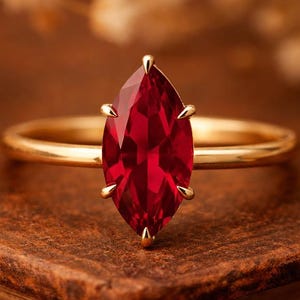 May include: A gold ring with a marquise-cut, deep red gemstone. The ring's band is thin and polished, and the gemstone is held in place by four prongs. The ring is set against a blurred background, highlighting the jewelry.