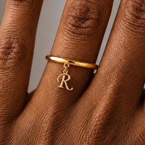 May include: A gold-toned ring featuring a thin, smooth band and a small, dangling charm in the shape of the capital letter R. The charm is attached to the band by a tiny circular link, creating a delicate and minimalist piece of jewellery.
