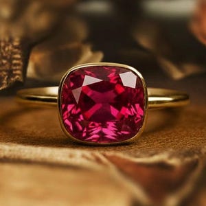 Peut inclure: Une bague en or ornée d'une grosse pierre précieuse rouge foncé de taille carrée. La pierre précieuse est sertie dans une lunette en or, et le corps de la bague est en or poli simple. La bague est présentée sur une surface en bois.