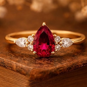 May include: A gold ring featuring a pear-shaped, deep red gemstone as the centerpiece, flanked by smaller white gemstones. The ring is set on a gold band and rests on a wooden surface, showcasing its elegant design.