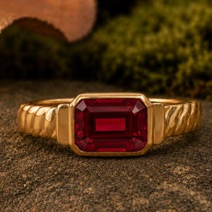 May include: A gold ring featuring a rectangular, emerald-cut deep red gemstone set in the center. The band of the ring displays a textured, ribbed pattern on both sides of the central stone setting, creating a distinct and structured appearance.