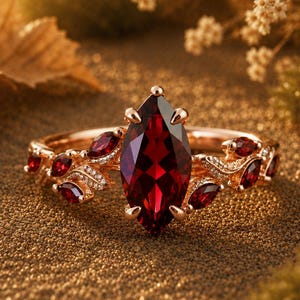 May include: A rose gold ring with a large, marquise-cut, deep red gemstone as the centerpiece. Smaller red gemstones and delicate detailing adorn the band. The ring is set against a textured, golden-brown surface, with blurred foliage in the background.