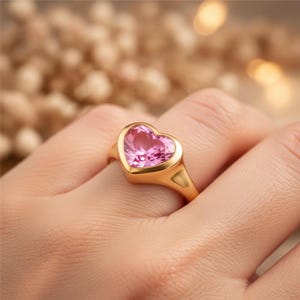 May include: A gold ring featuring a heart-shaped pink gemstone. The ring has a smooth, polished band and a bezel setting that encases the gemstone. The heart-shaped stone is a vibrant pink color.