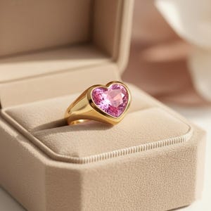 May include: A gold ring with a large, heart-shaped pink gemstone. The ring is set in a thick, polished gold band and is displayed in a beige jewelry box. The gemstone has multiple facets, reflecting light.