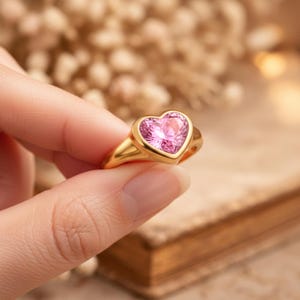 May include: A gold ring featuring a heart-shaped pink gemstone. The ring is held between fingers, showcasing its polished gold band and the vibrant pink hue of the heart-shaped stone. The ring is a piece of jewelry.