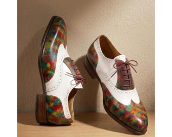 May include: A pair of two-tone leather dress shoes. The shoes have a white upper with colorful, patterned accents in red, green, and yellow. Brown laces and soles complete the design. The shoes are displayed on a wooden surface.
