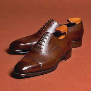 Peut inclure: Une paire de chaussures de ville en cuir marron poli, avec des lacets noirs et des embauchoirs en bois. Les chaussures ont un design classique à bout rapporté, sur fond brun-rouge.