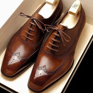 May include: A pair of brown leather dress shoes with intricate brogue detailing on the toe and sides. The shoes have a classic lace-up design and are placed in a wooden box with shoe trees inside.