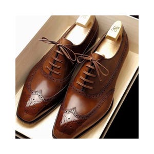 May include: A pair of brown leather dress shoes with brogue detailing and a lace-up closure. The shoes are displayed in a wooden box with shoe trees inserted. The leather has a polished, glossy finish.