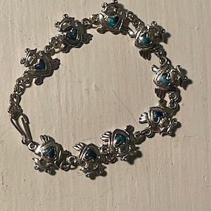 May include: Silver-toned bracelet featuring a series of stylized frog charms. Each frog has a heart-shaped blue center. The bracelet is connected by small chain links and has a clasp closure. The bracelet is displayed on a white surface.