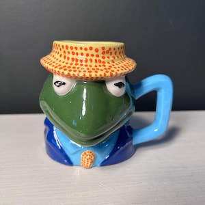 May include: A ceramic frog-shaped mug with a green head, blue suit, and a yellow hat with orange polka dots. The mug has a blue handle and is set against a white surface.
