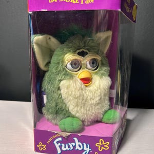 May include: A green and cream-colored electronic Furby toy inside its original purple packaging. The box features the text 'The more you PLAY with me the MORE I do!' and 'Furby'. The toy has large blue eyes, long eyelashes, and a yellow beak.