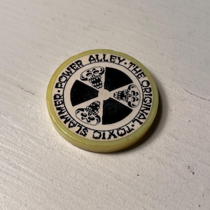 May include: A round, off-white token with the text "POWER ALLEY - THE ORIGINAL - TOXIC SLAMMER" encircling a black and white graphic of a skull and crossbones design. The token has a yellow-green edge.