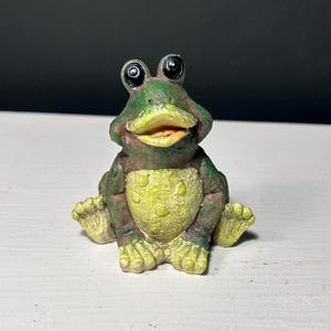 May include: A small, seated frog figurine with a textured green and brown body, a pale yellow belly with raised spots, and large, prominent black eyes. The frog has an open mouth, visible webbed feet, and a detailed, weathered surface finish.