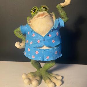 May include: A whimsical frog figurine with a light green body and a light blue outfit adorned with pink flower accents. The frog has large, expressive eyes and is posed with one arm raised. The figurine is a decorative item.