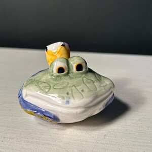 May include: A ceramic frog figurine with a green body, white belly, and blue and yellow accents. The frog has large, black-pupiled eyes and a small yellow bird perched on its head. The figurine is likely a decorative item.