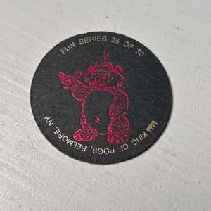 May include: A black, circular Pog Slammer featuring a red cartoon character with the text "FUN SERIES 25 OF 30" and "KING OF POGS, BELMORE, NY". The character is holding a drink.