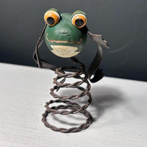 May include: A decorative frog figurine featuring a round green wooden head with large orange and black eyes, a painted mouth, and thin metal arms. The body consists of a coiled, twisted dark brown metal spring that serves as a base for the figure.