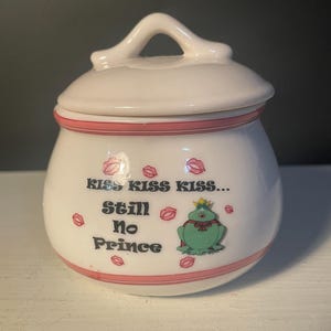 May include: White ceramic jar with a lid and handle, featuring pink accents and the text "Kiss Kiss Kiss... Still No Prince." Adorned with cartoon lips and a frog wearing a crown. A decorative kitchen or bathroom accessory.
