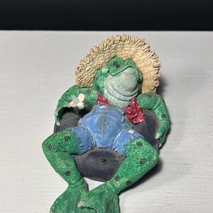 May include: A green frog figurine wearing a straw hat, blue denim overalls, and a red polka-dot neckerchief. The frog is lounging in a black inner tube, with its webbed feet extended forward and its arms resting on the sides of the tube.