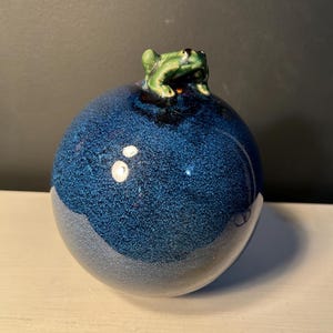 May include: A round, blue ceramic decorative object with a textured surface. A small green frog figurine sits atop the sphere. The frog has black eyes and is detailed with green and black paint.