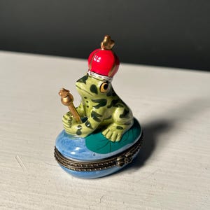 May include: A ceramic frog figurine wearing a red crown and holding a golden scepter. The frog is green with dark spots and sits on a blue and green lily pad. The figurine is a decorative trinket box.