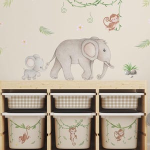 May include: A wooden storage unit with six compartments, three with checkered bins and three with jungle-themed bins featuring monkeys. The wall behind has watercolor illustrations of elephants, monkeys, and foliage, creating a nursery theme.