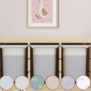 May include: Three white storage bins with a light blue scalloped design and pink bow accents. The bins are set in a wooden frame, with a framed illustration of a goose above. A selection of colored circles with bow designs are in the foreground.