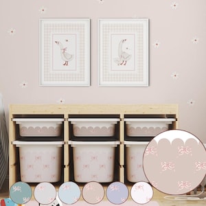 Pode incluir: Uma cena de quarto de criança com duas ilustrações de gansos emolduradas com laços rosa, acima de caixas de armazenamento. As caixas e a parede têm um padrão de laços correspondente. O quarto é decorado com decalques de margaridas e uma unidade de armazenamento de madeira.