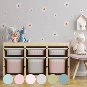 Può includere: Un mobile contenitore in legno con sei contenitori, tre grigi e tre rosa, si trova sotto un muro grigio decorato con decalcomanie a forma di margherita. Peluche, tra cui un elefante, un rinoceronte e una volpe, sono in cima. Una mensola bianca contiene una statuina di gatto.