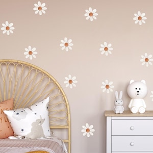 Op de afbeelding: Een slaapkamer met madeliefjes muurstickers, een rotan hoofdbord en decoratieve kussens. Een witte ladekast bevat een beer-vormige lamp en een konijn figuur. Het kleurenschema is zacht, met beige, witte en oranje accenten.