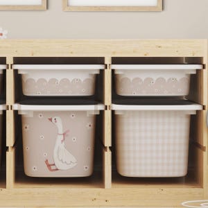 Könnte beinhalten: Ein Aufbewahrungsmöbel aus hellem Holz mit vier weißen Kunststoffbehältern. Die Behälter zeigen beige Muster, darunter eine weiße Gänseillustration, kleine Gänseblümchen, gewellte Ränder und ein hellrosa Vichy-Karomuster auf den Fronten.