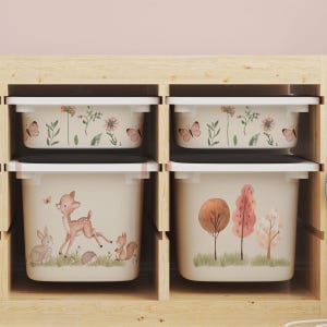 Könnte beinhalten: Vier beige Aufbewahrungsboxen mit weißen Deckeln sind in einem Holzregal angeordnet. Die oberen Boxen weisen ein Blumen- und Schmetterlingsdesign auf. Die unteren Boxen zeigen Waldszenen mit einem Reh, einem Eichhörnchen und Bäumen.