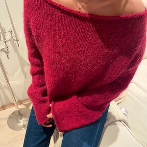 Könnte beinhalten: Ein leuchtend roter Off-Shoulder-Pullover. Der Pullover hat lange Ärmel und einen weiten Ausschnitt. Das Material scheint ein weiches, strukturiertes Strick zu sein. Der Pullover wird mit einer blauen Jeans kombiniert. Eine goldene Halskette ist sichtbar.